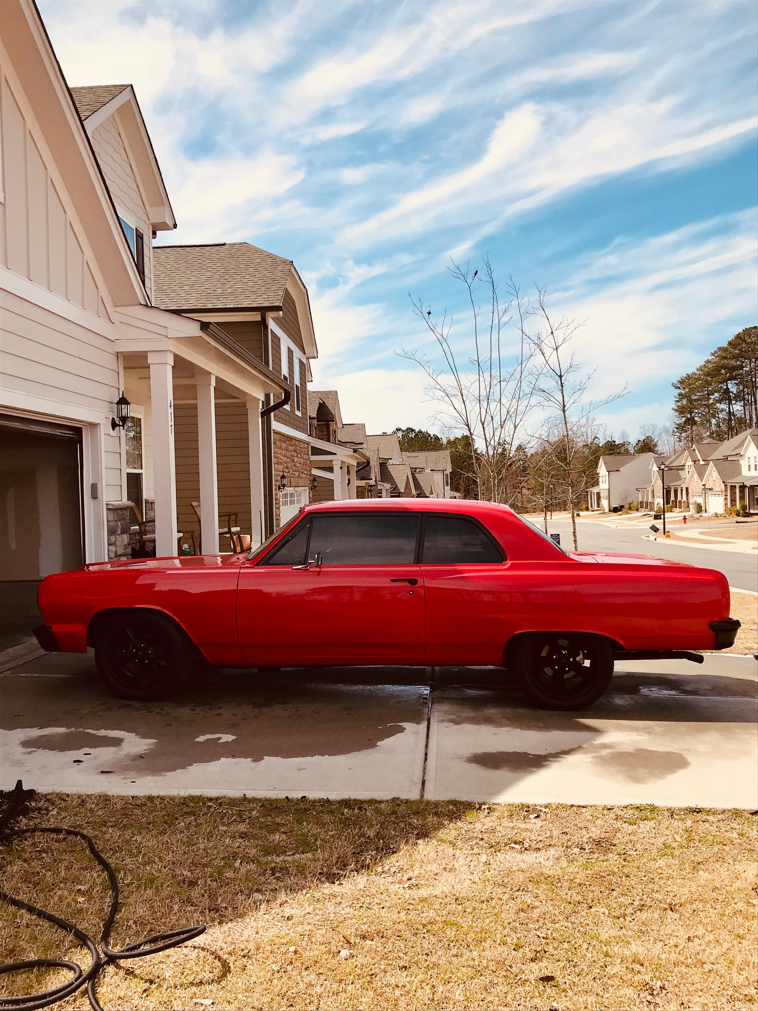 Bright red classic muscle car with black wheels parked in a sunny suburban driveway.
